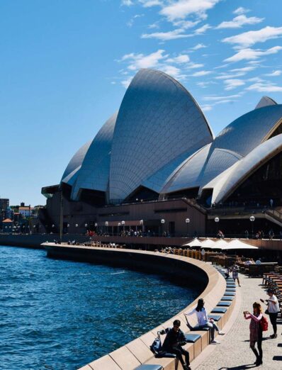 ydney Opera House with its iconic white sails overlooking the blue waters of Sydney Harbour, under a bright blue sky with visitors walking along the waterfront promenade.