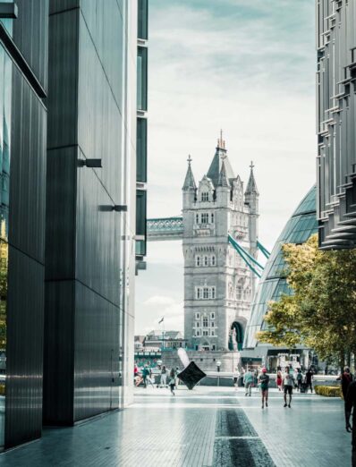 View of London’s Tower Bridge framed between modern glass buildings near City Hall, with pedestrians walking along the South Bank on a bright day.